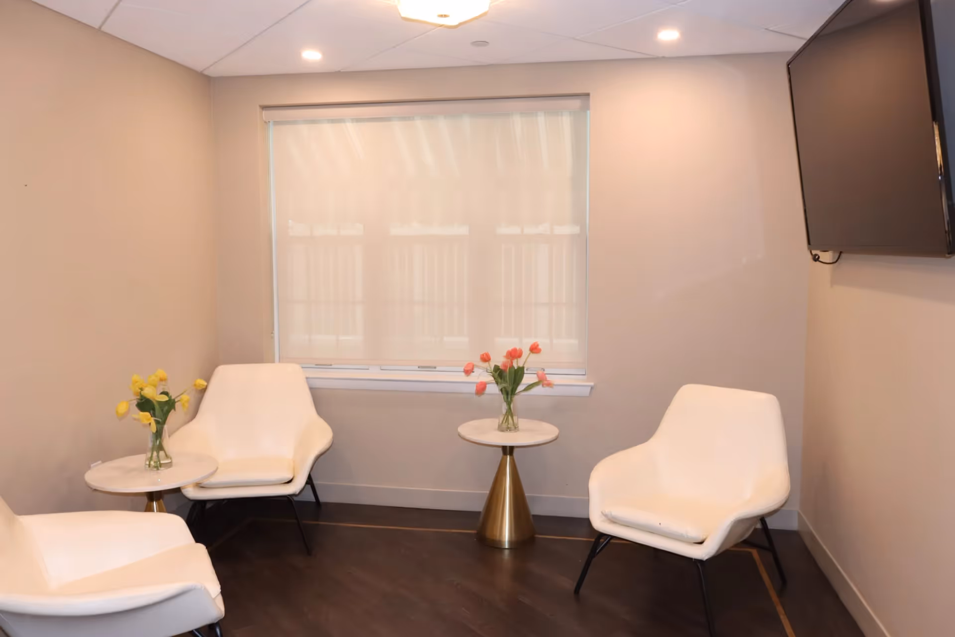 A small, modern lounge area with white chairs, round tables with flower vases, a window with blinds, and a wall-mounted TV.