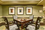 A cozy interior room with a wooden table and four cushioned chairs arranged around it. The walls are painted green and decorated with three framed pictures of doors. The room has a warm and inviting atmosphere with soft lighting.