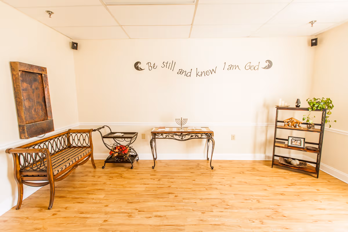 A bright room with light wooden flooring and cream-colored walls. The room features a wooden bench with decorative metal armrests on the left, a small black metal cart with red flowers underneath, a decorative table with a menorah and books in the center, and a black shelving unit with plants, framed photos, and decorative items on the right. Above the table, a wall decal reads 'Be still and know I am God' with crescent moon designs on either side.