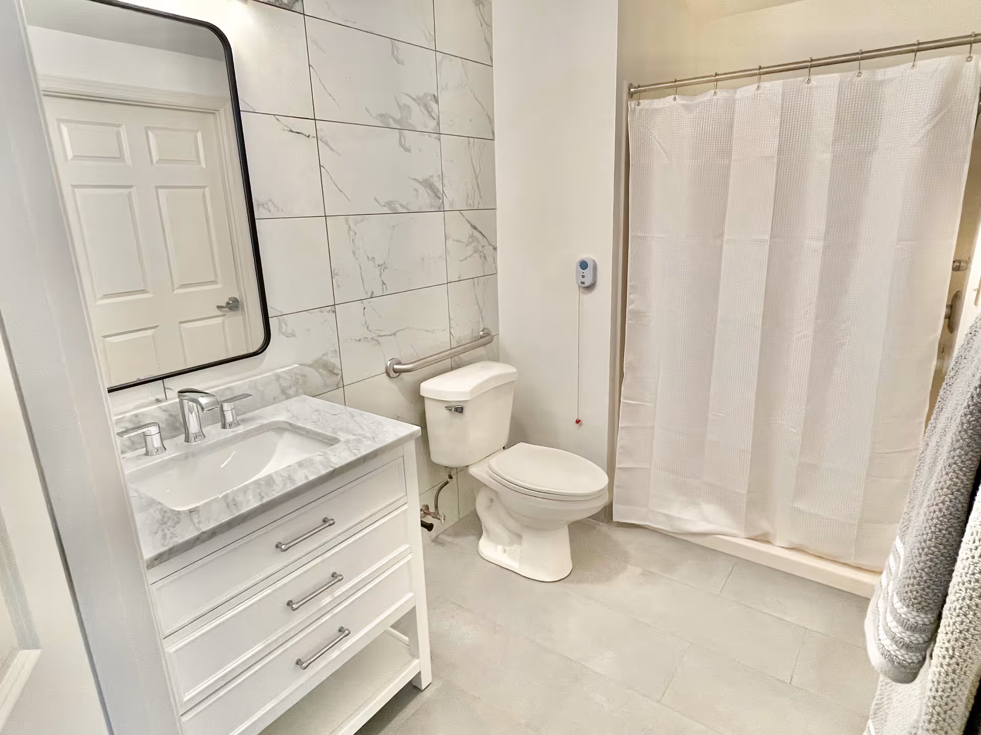 A clean and modern bathroom with a white vanity featuring a marble countertop and a rectangular mirror above it. There is a white toilet next to the vanity and a shower with a white curtain. The walls have large marble-patterned tiles and there is a grab bar installed next to the toilet. A pull cord emergency alert is mounted on the wall near the shower.