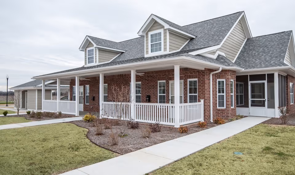 Exterior view of a single-story senior living facility building with red brick walls, beige siding, and a gray shingled roof. The building features a covered porch with white railings and several windows. There is a concrete walkway leading to the entrance, surrounded by grass and landscaped areas with small shrubs and plants.