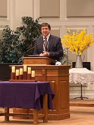 A man in a suit standing behind a wooden podium speaking into a microphone in a room with paneled walls. In front of him is a table covered with a purple cloth holding several candles. To the right, there is a table with a white lace cloth and a large vase filled with yellow flowers. Green plants are visible in the background.