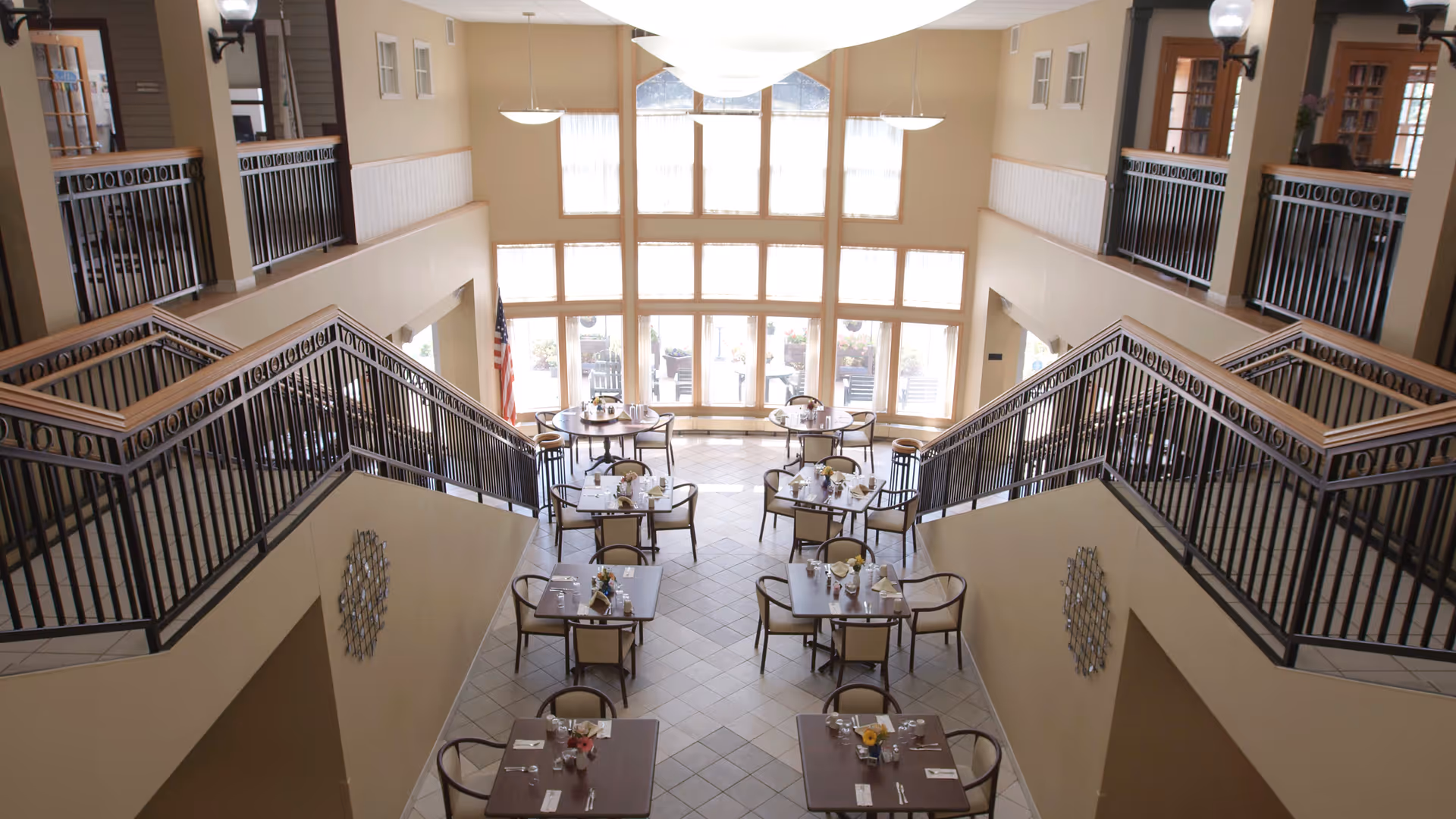 A bright dining area with multiple tables set for meals, each with chairs around them. The room has large windows letting in natural light, and there are railings on both sides of a staircase leading down to the dining space. The walls are beige, and there are decorative wall hangings and an American flag near the windows.