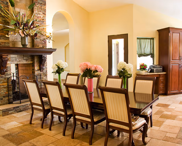 A dining room with a long dark wooden table surrounded by eight upholstered chairs. The table is decorated with vases of pink and white flowers. To the left, there is a stone fireplace with a large floral arrangement on the mantel. The room has a tiled floor, cream-colored walls, and a wooden cabinet in the background near a window with a green curtain.