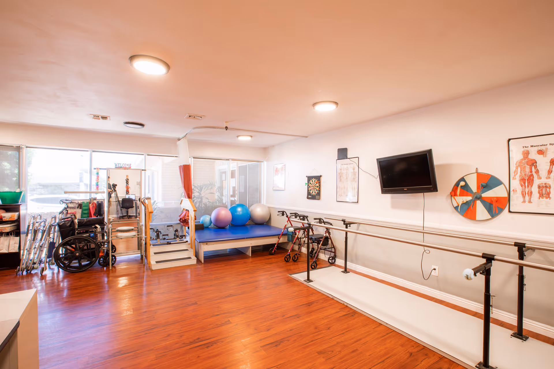 A rehabilitation or physical therapy room with wooden flooring, parallel bars for walking exercises, exercise balls on a blue platform, walkers, a wheelchair, and various medical and exercise equipment. The room has large windows letting in natural light, a wall-mounted TV, anatomical charts, and a dartboard.