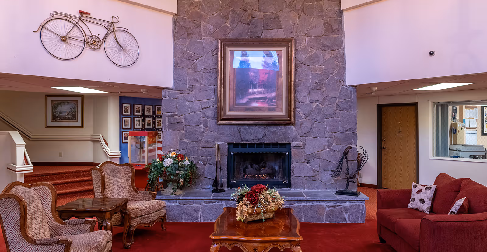 A cozy senior living community lounge area featuring a large stone fireplace with a framed painting above it. In front of the fireplace is a wooden coffee table with a floral arrangement. To the left, there are two upholstered armchairs with a small wooden side table between them. To the right, a red sofa with patterned pillows is visible. The room has red carpeting and a decorative metal bicycle mounted on the wall above the fireplace. There are stairs leading to another area and a popcorn machine in the background.
