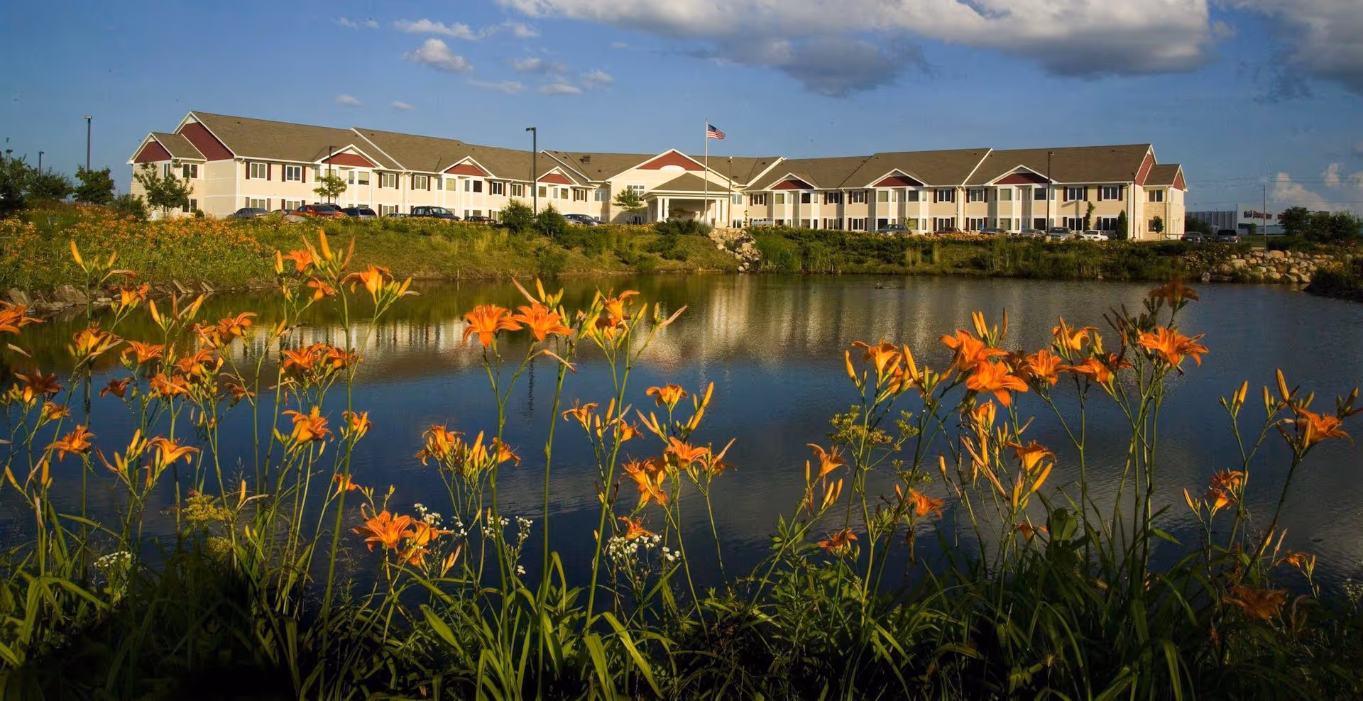 A long senior living facility building sits across a pond with orange flowers in the foreground under a partly cloudy sky.
