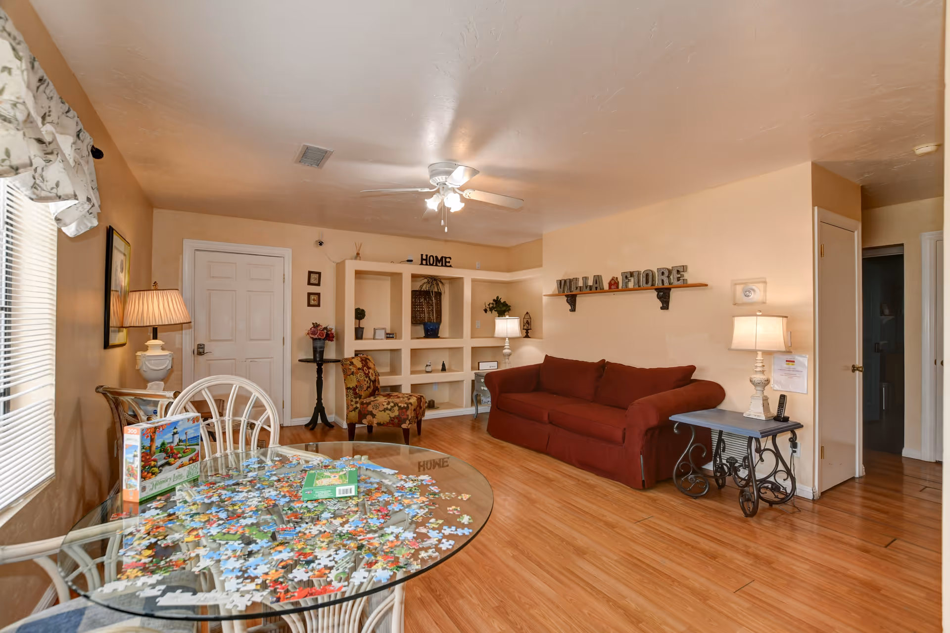 A cozy living room with a red sofa, a floral armchair, and a glass table with a partially completed jigsaw puzzle. The room has wooden flooring, beige walls, a ceiling fan with lights, and built-in shelves decorated with plants and ornaments. There is a lamp on a side table next to the sofa and a window with blinds and a floral valance.