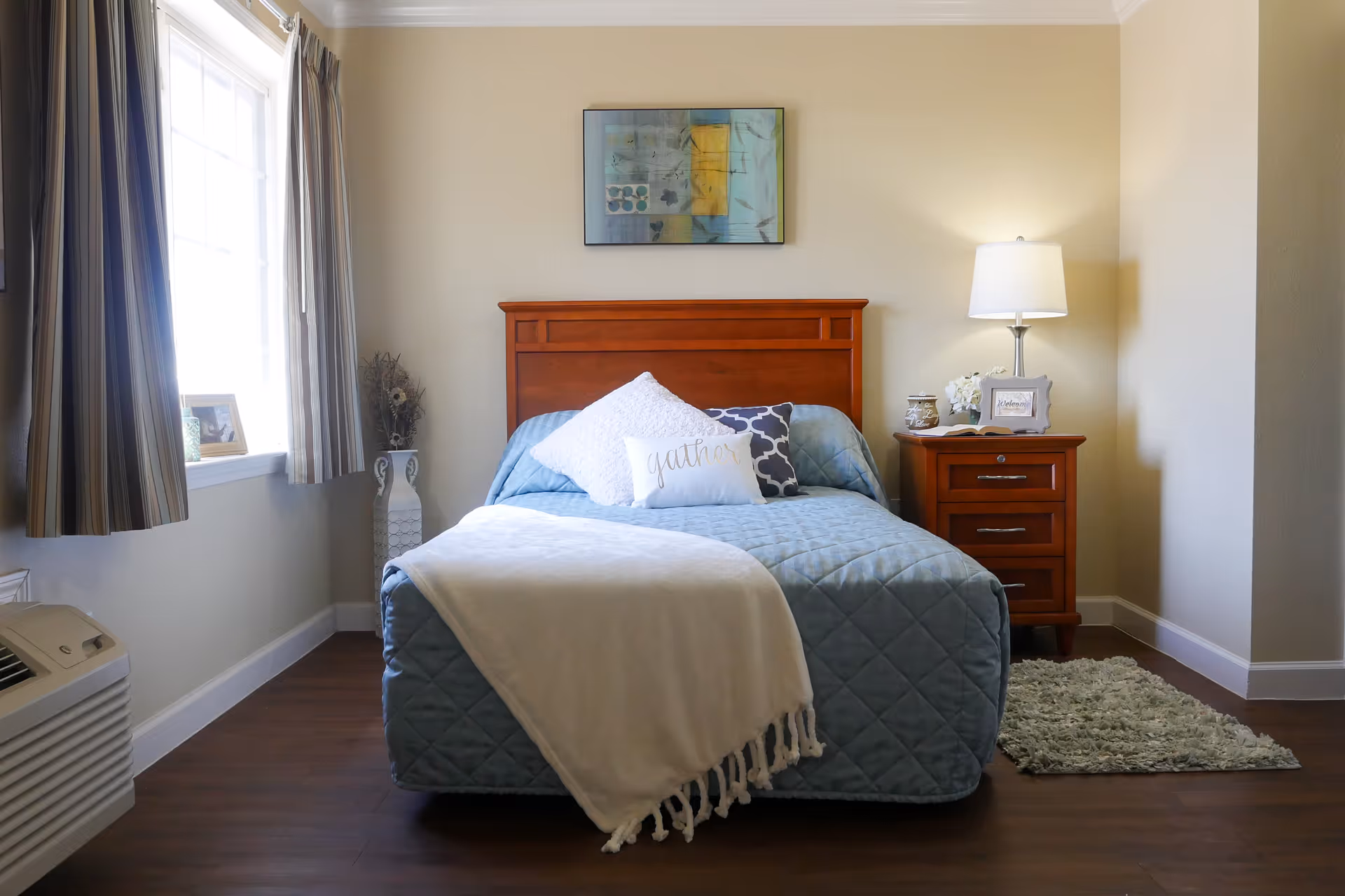 A bright, neatly made bedroom featuring a bed with a light-blue quilt and wooden headboard, a nightstand with lamp, a window with striped curtains, and a small rug.