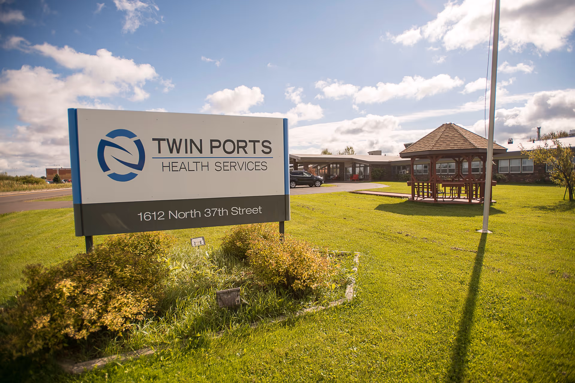 Outdoor view of a Twin Ports Health Services sign on a grassy lawn with the facility building and a gazebo in the background.