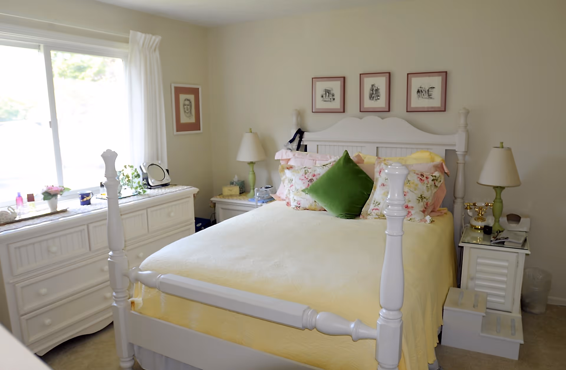 A cozy bedroom with a white wooden bed frame and a yellow bedspread. The bed is decorated with floral pillows and a green accent pillow. There are two white nightstands on either side of the bed, each with a lamp. A white dresser with various items on top is positioned under a large window with white curtains. Three framed pictures hang on the wall above the bed.