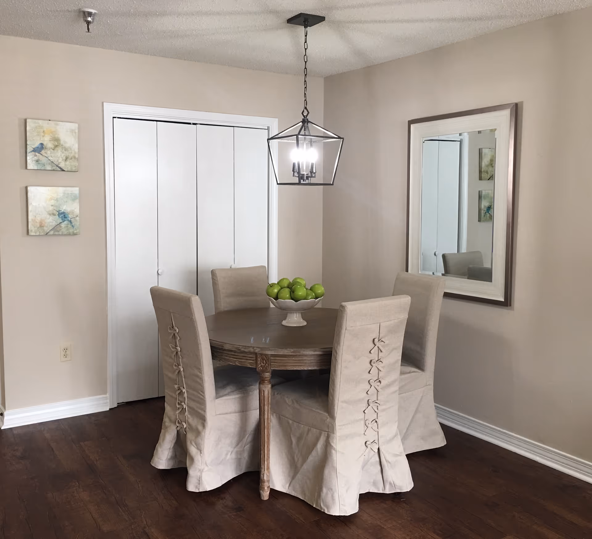 Small dining area with a round wooden table, four beige slipcovered chairs, a hanging light fixture, a wall mirror and a bowl of green apples.