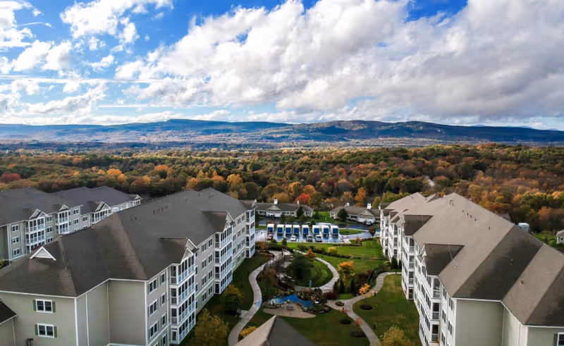 Aerial view of Woodland Pond At New Paltz senior living facility showing multiple large residential buildings surrounding a landscaped courtyard with walking paths, a small pond, and a pool area with lounge chairs. The background features a vast expanse of forested hills under a partly cloudy sky.