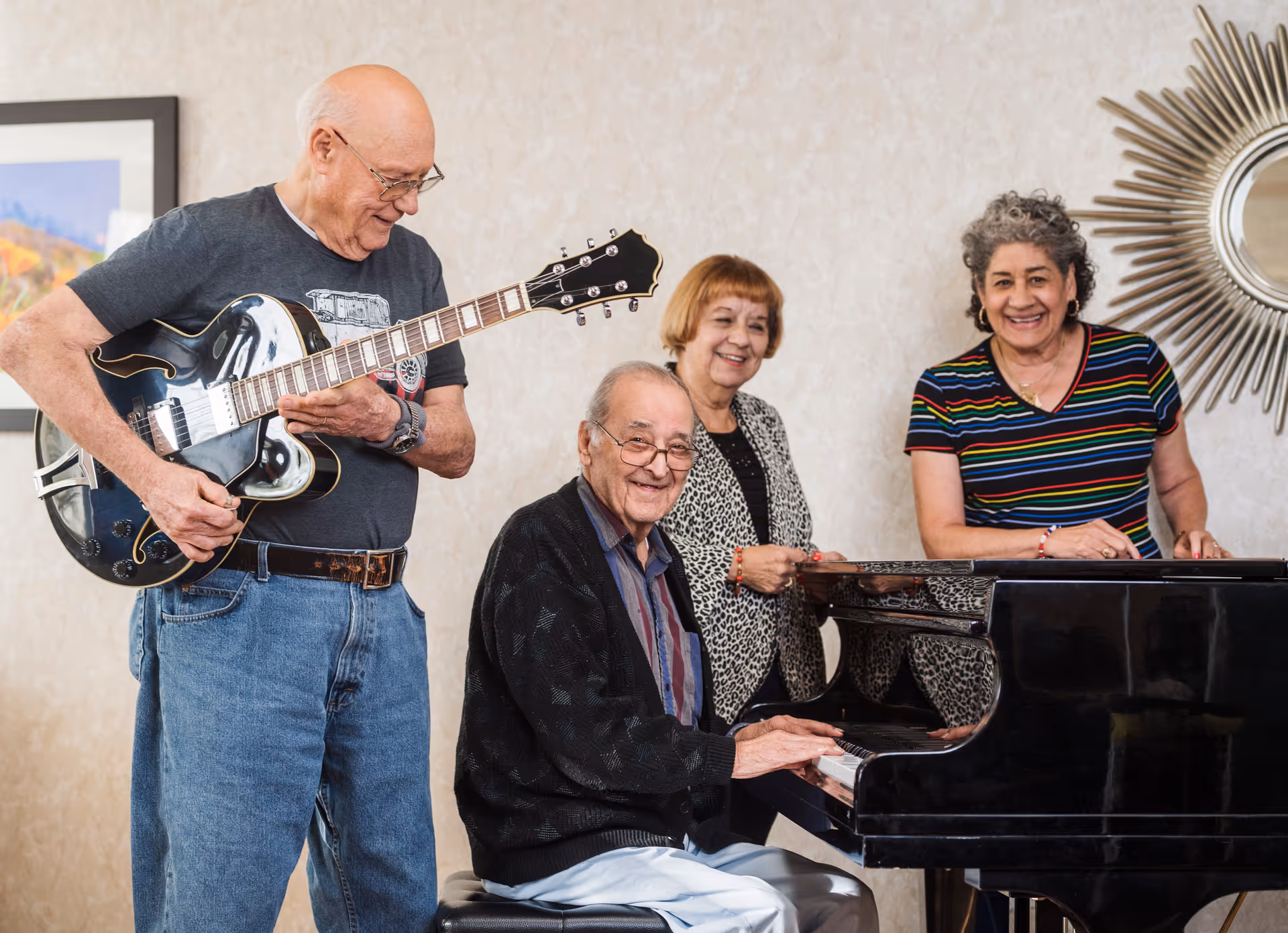 Four elderly people enjoying music together indoors. One man is playing an electric guitar, another man is seated playing a grand piano, and two women are standing nearby smiling. The room has light-colored walls, a framed picture, and a decorative mirror.