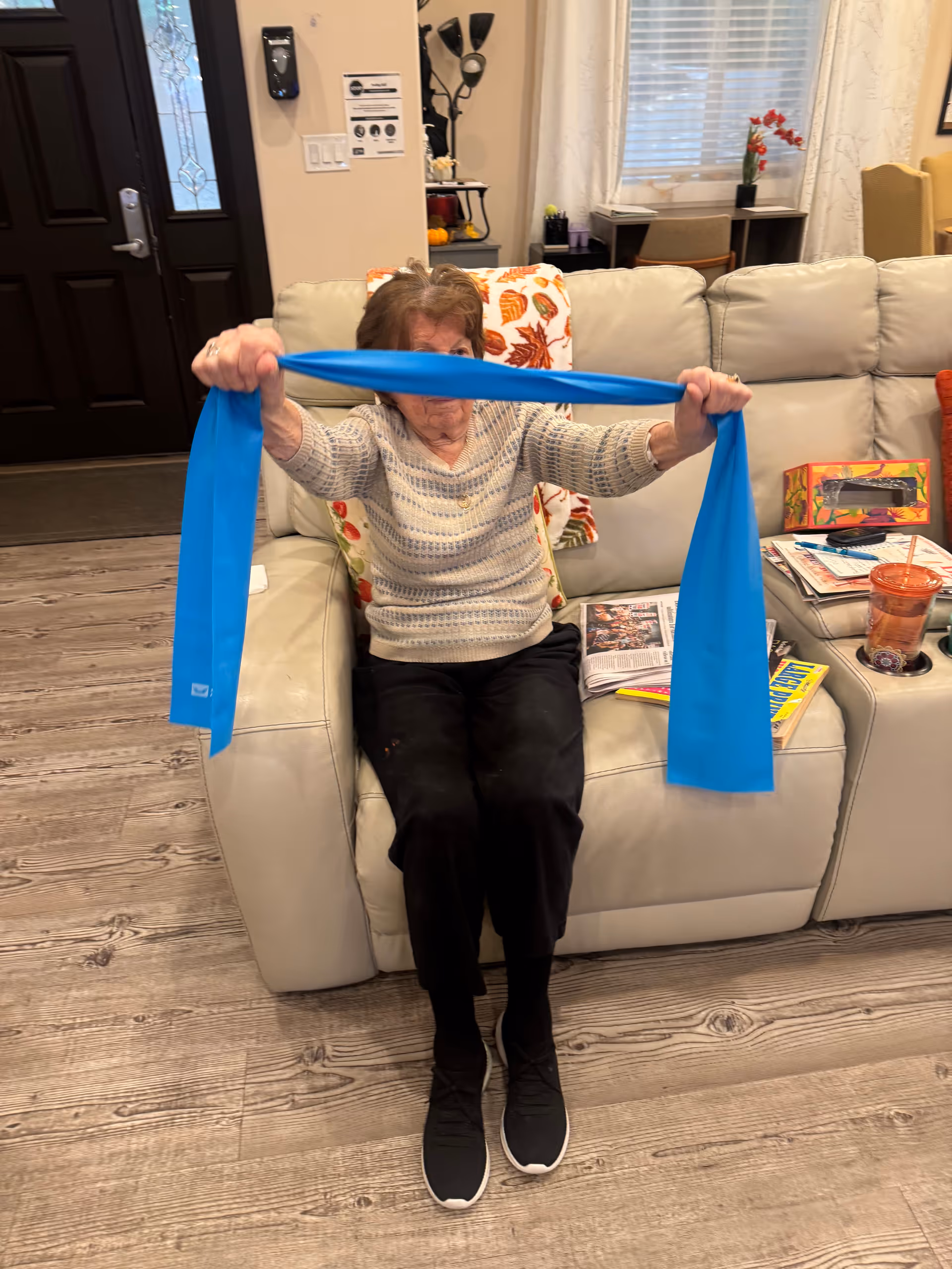 An elderly woman sitting on a light-colored leather couch in a living room, holding and stretching a blue exercise resistance band with both hands. The room has wooden flooring, a dark front door, and a side table with various items including a cup and magazines. There is a decorative pillow and a blanket with autumn leaf patterns on the couch.