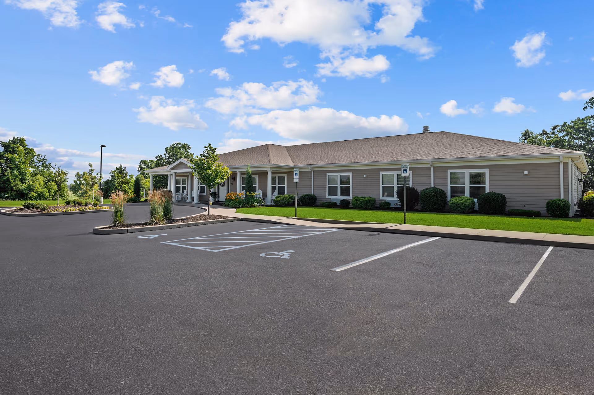 Exterior view of a single-story senior living facility building with a beige facade, multiple windows, and a covered entrance. The foreground shows a paved parking lot with designated handicap parking spaces and landscaped areas with trees and shrubs. The sky is blue with scattered clouds.