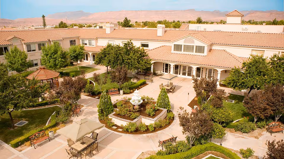 Aerial view of a senior living facility courtyard with a central fountain surrounded by landscaped gardens, trees, benches, and outdoor seating areas with tables and umbrellas. The building has a tiled roof and large windows, with mountains visible in the background under a clear sky.