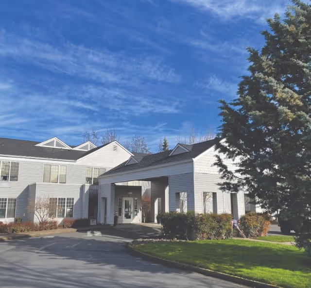 Exterior view of a senior living facility building with white siding and a covered entrance. There is a large tree on the right side and a clear blue sky with some clouds above. The driveway and some landscaping with bushes and grass are visible in the foreground.