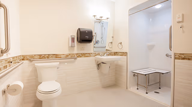 Accessible senior living bathroom with a toilet, wall-mounted sink, grab bars, soap and paper towel dispensers, and a roll-in shower with a folding bench.