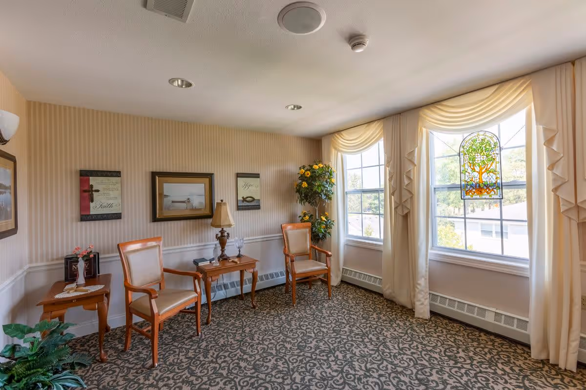 A cozy sitting area in a senior living facility with two wooden armchairs with beige cushions, two small wooden side tables, a table lamp, framed artwork on the wall, a tall potted plant with yellow flowers, and large windows with cream-colored curtains and a stained glass decoration hanging in the center window.