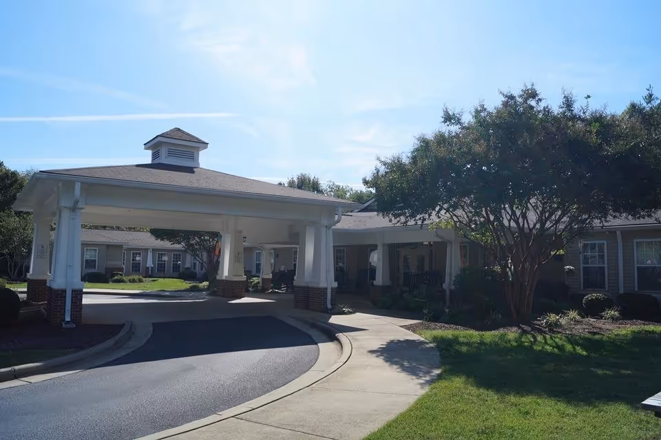 Exterior view of Somerset Court of Mocksville showing a covered driveway entrance with white columns and brick bases, surrounded by green lawns, trees, and shrubs under a clear blue sky.