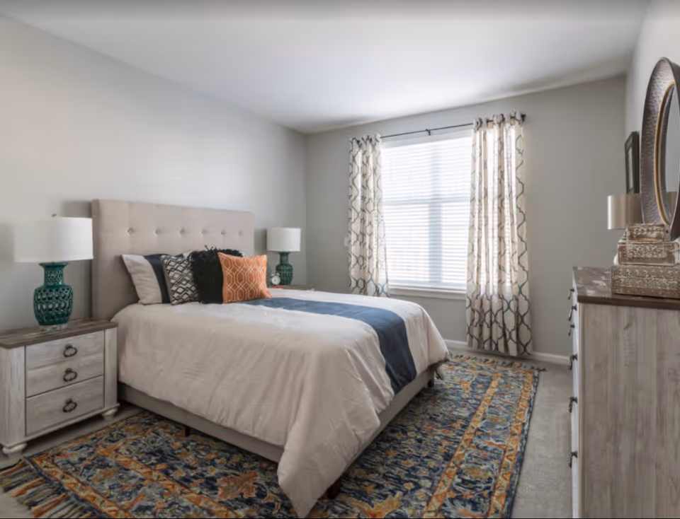 A bright, modern bedroom with a tufted headboard bed, nightstands with lamps, patterned rug, and a dresser by a window with curtains.