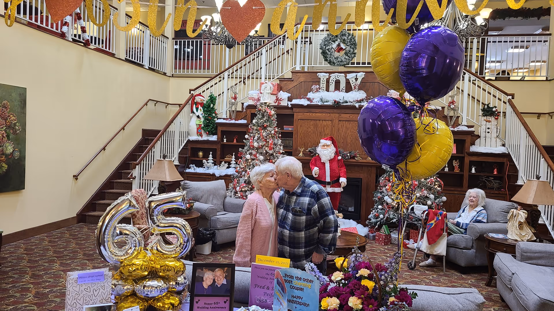 An elderly couple kissing in the center of a decorated living room celebrating their 65th wedding anniversary. The room features festive holiday decorations including Christmas trees, a Santa Claus figure, and wreaths. Large silver and gold balloons shaped as the number 65 are displayed on a table with framed photos and congratulatory cards. Another elderly woman is seated on a couch in the background. The space has a staircase with white railings and warm lighting.