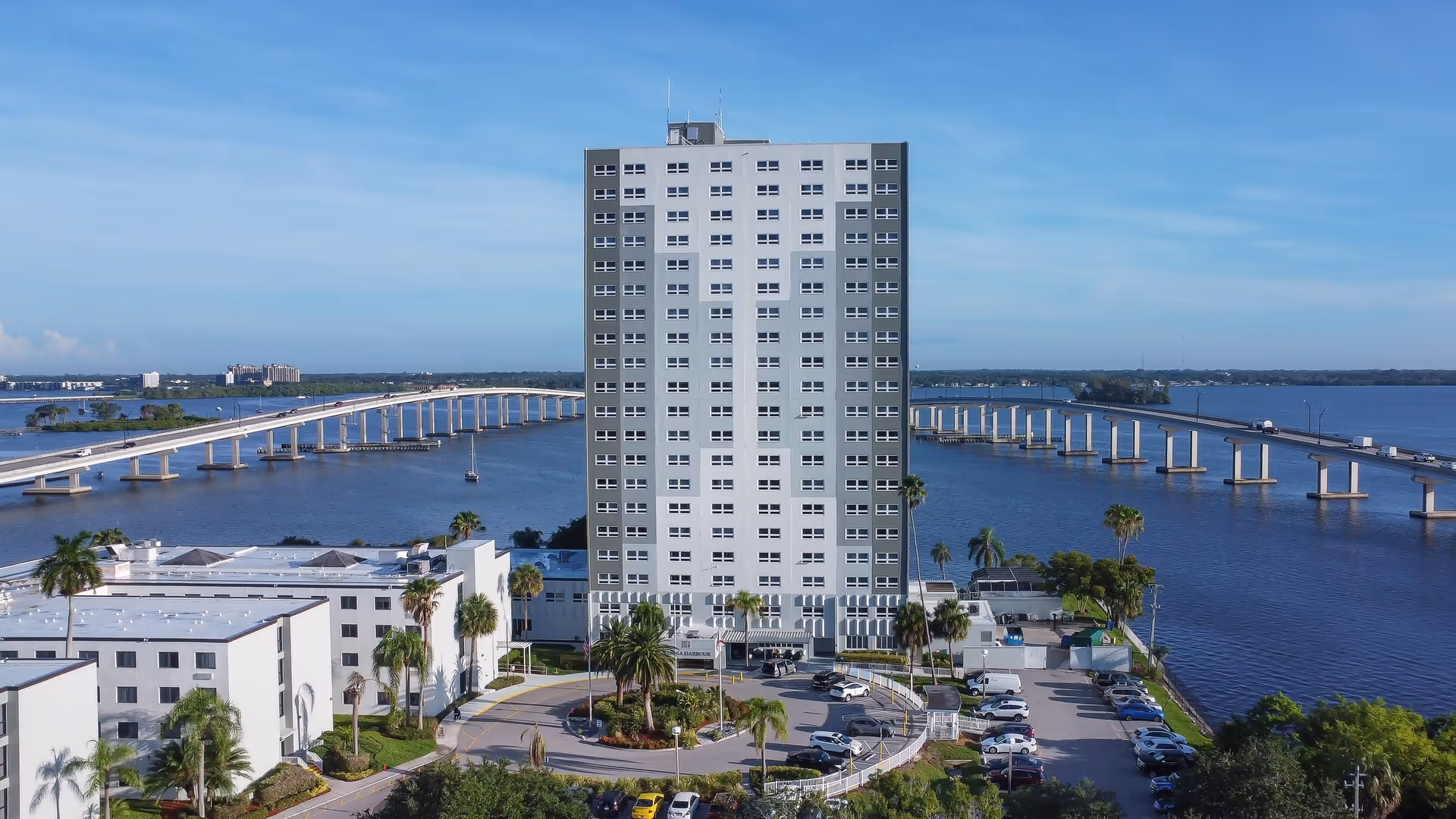 Tall multi-story building named Calusa Harbour situated near a large body of water with a long bridge extending across the water in the background. The building is surrounded by palm trees, parking lots, and smaller white buildings under a clear blue sky.