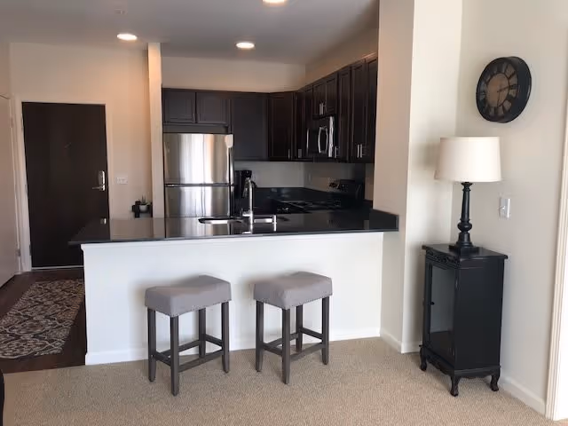 Modern kitchen area with dark wood cabinets, stainless steel refrigerator, microwave, and stove. A black countertop bar with two gray cushioned stools is in the foreground. To the right, there is a small black cabinet with a lamp on top and a round wall clock above it. The entrance door is visible in the background.