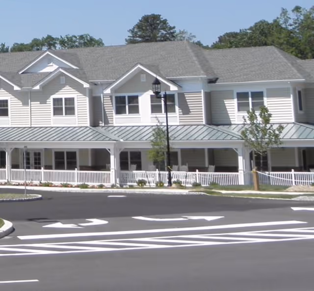 Exterior view of a two-story senior living facility building with beige siding, white trim, a covered porch with white railings, and a paved driveway with directional arrows in front. Trees and landscaping are visible around the building.