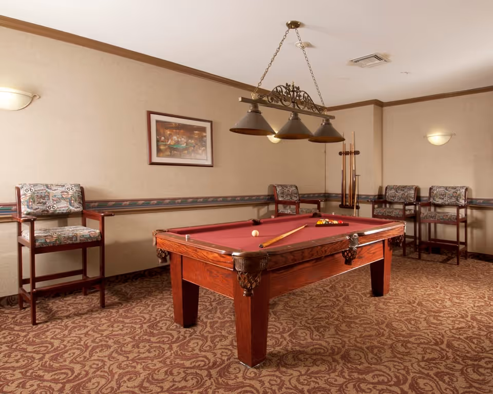 A game room with a red felt pool table in the center, a pool cue and balls on the table, four patterned cushioned chairs along the walls, a framed picture on the beige wall, and a hanging light fixture with three lamps above the pool table.