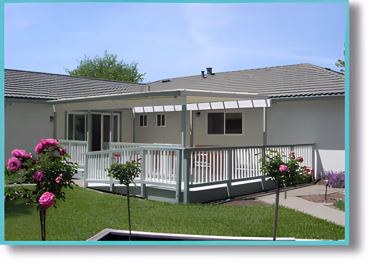 Outdoor view of a single-story building with a covered porch and white railing. The area is surrounded by green grass and blooming pink flowers under a clear blue sky.