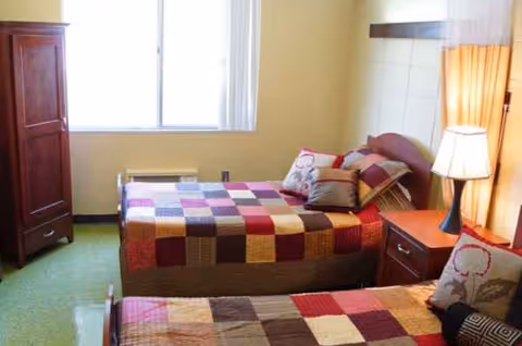 Two twin beds with patchwork quilts in a sunny senior living bedroom.