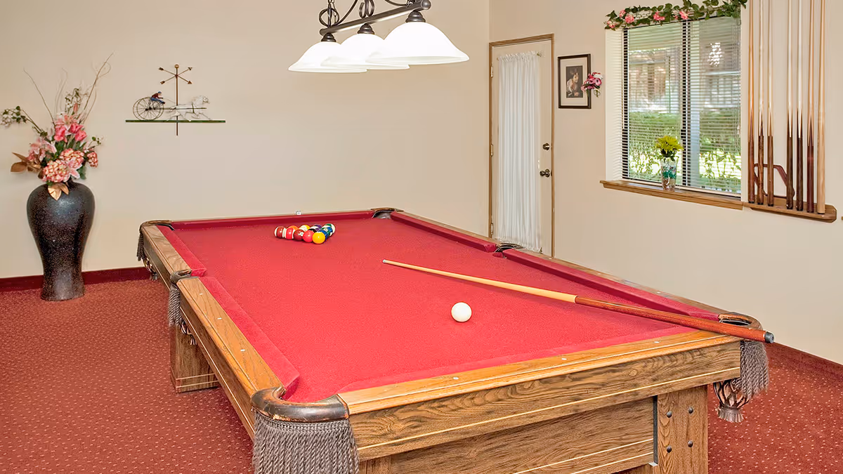 A room with a red-felt pool table set up for a game, with a cue stick resting on the table. The room has beige walls, a window with blinds, a door with a curtain, a large vase with flowers, and a wall-mounted decorative piece. A ceiling light fixture with three lamps hangs above the pool table.
