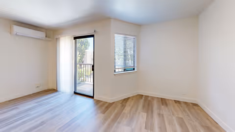 Empty room with light wood flooring, white walls, a sliding glass door leading to a balcony, a window with blinds, and a wall-mounted air conditioning unit.