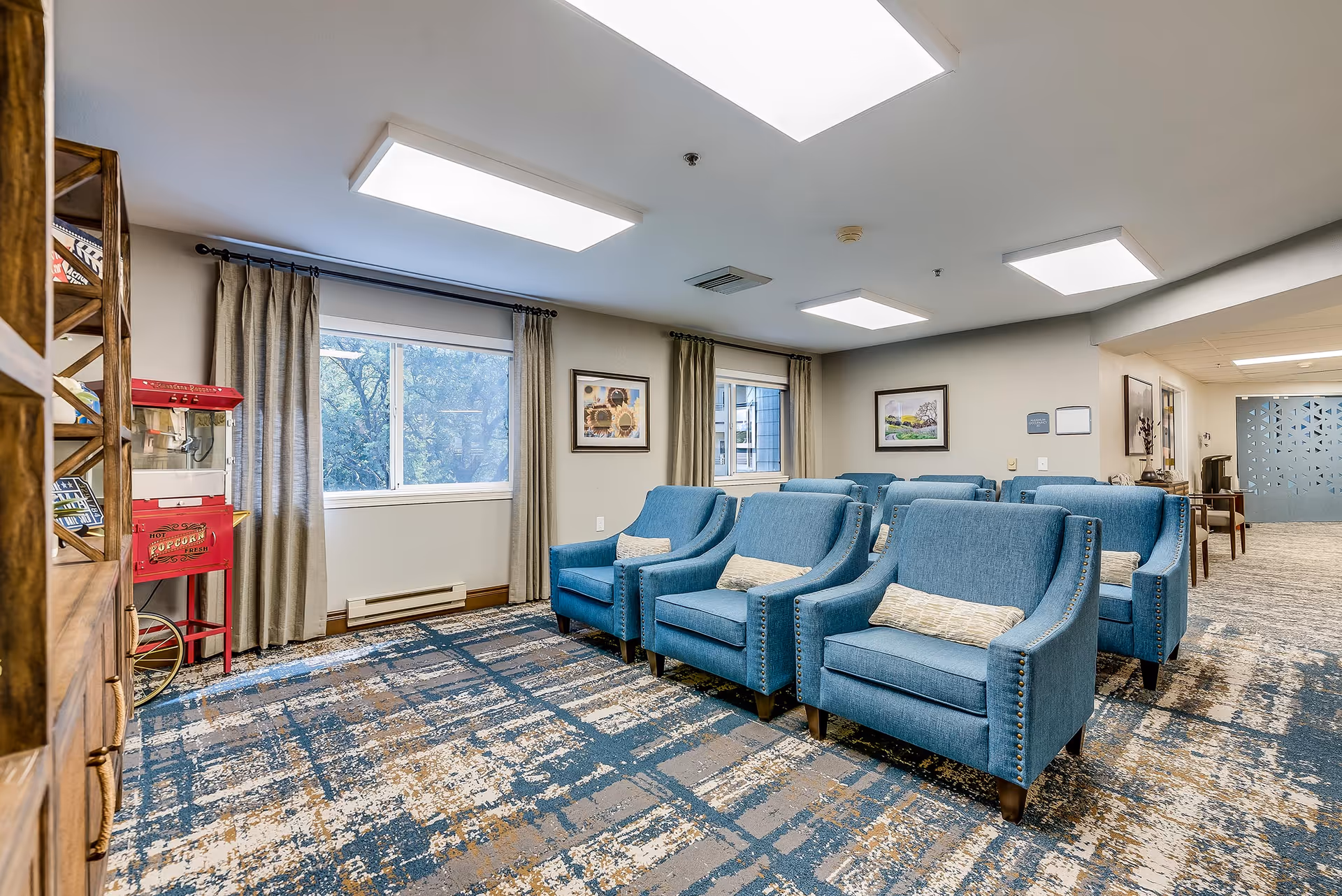 A cozy seating area in a senior living facility with two rows of blue upholstered armchairs, each with a small decorative pillow. The room has large windows with beige curtains, framed artwork on the walls, and a patterned carpet in shades of blue, beige, and brown. A red popcorn machine is visible near a wooden shelving unit on the left side of the image.