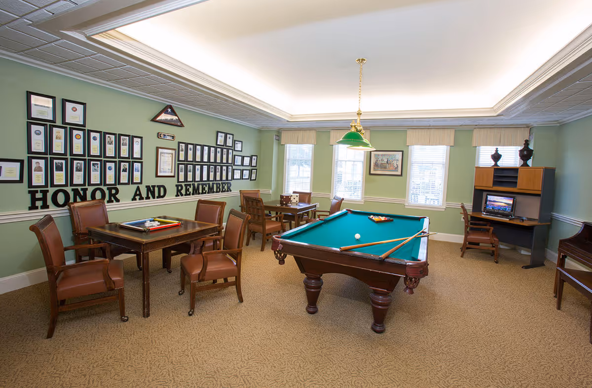 Well-lit recreation room with a pool table, card tables and chairs and a wall display reading "HONOR AND REMEMBER".