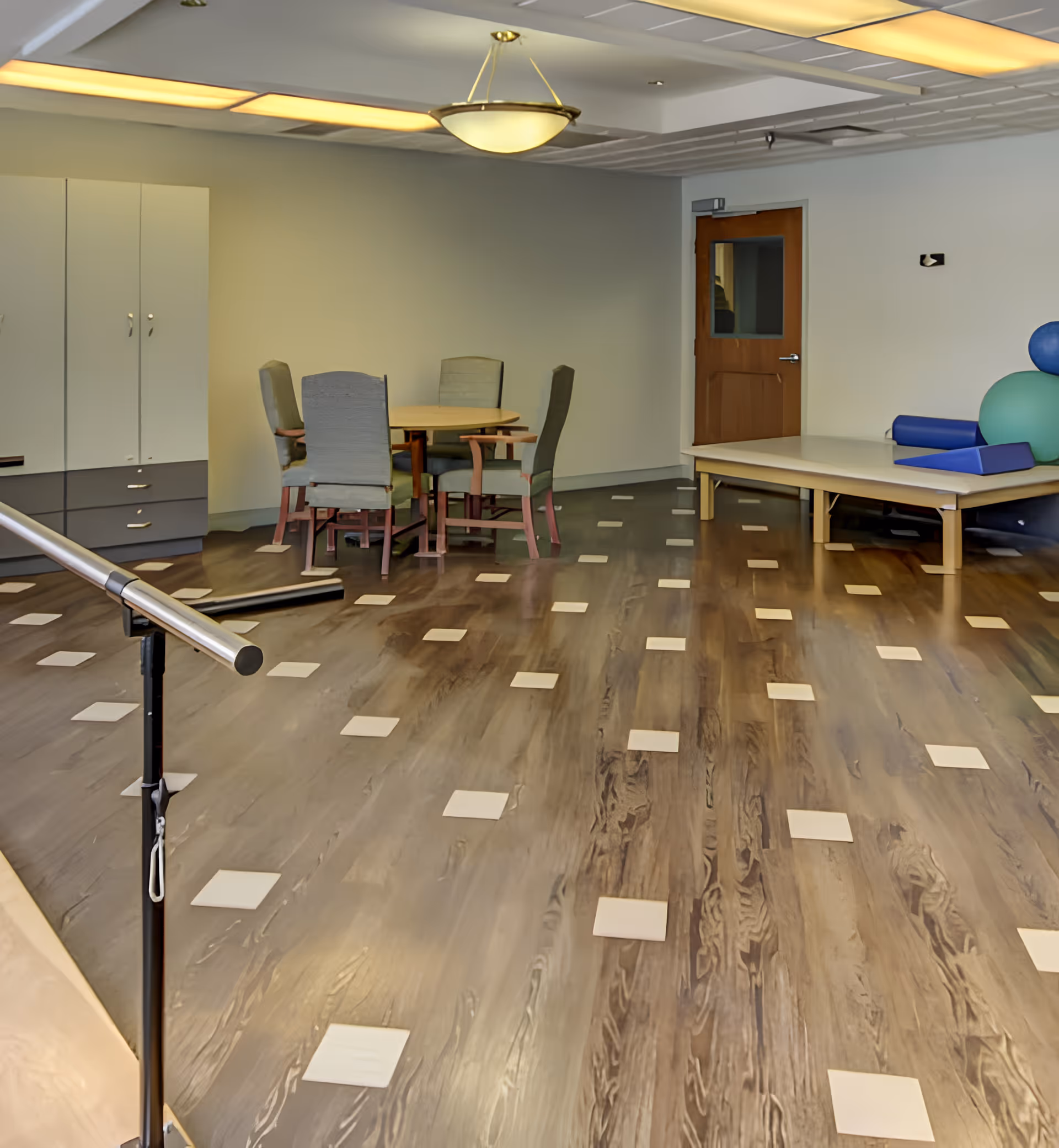 A spacious room with wooden flooring featuring square tile accents, a round table with four chairs, a wooden door with a window, a cabinet, and a therapy or exercise area with a padded table and large exercise balls. The ceiling has recessed lighting and a hanging light fixture.