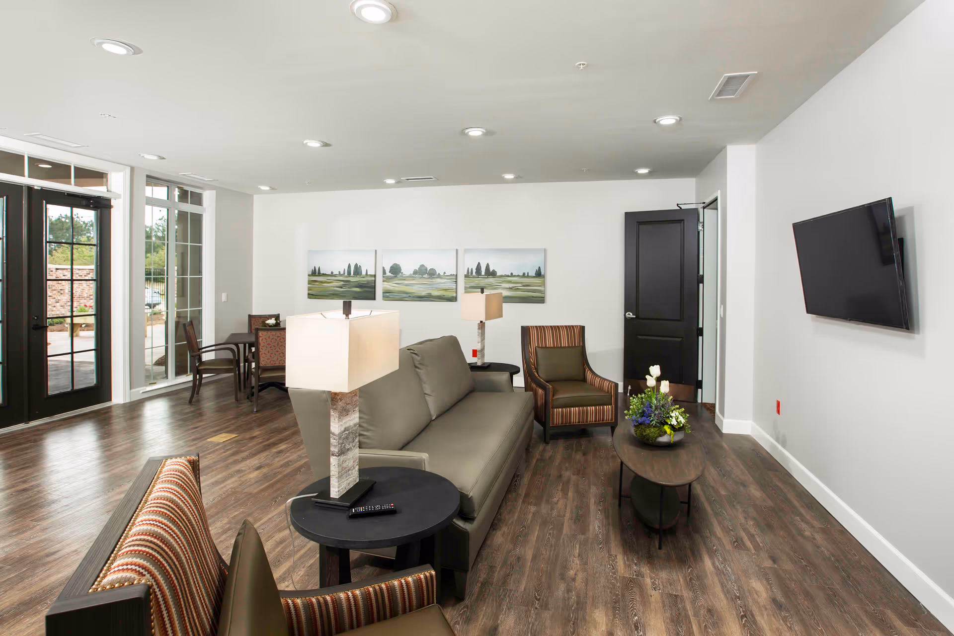 A modern living room in a retirement community with a gray sofa, two striped armchairs, a wooden coffee table with a flower arrangement, and a wall-mounted flat-screen TV. The room has wood flooring, white walls, recessed ceiling lights, and large windows and glass doors leading outside. There are three landscape paintings on the wall and a small dining table with chairs in the corner.