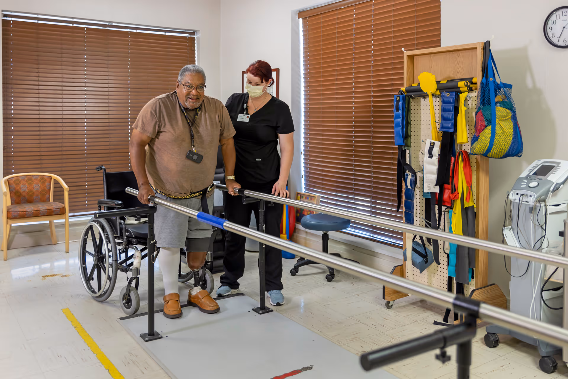 A man with a prosthetic leg practices walking with parallel bars in a rehabilitation room, assisted by a healthcare worker wearing a mask. A wheelchair and various therapy equipment are visible in the background.