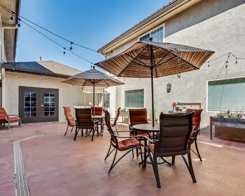 Outdoor courtyard patio with round tables, chairs, and striped umbrellas between buildings under string lights.