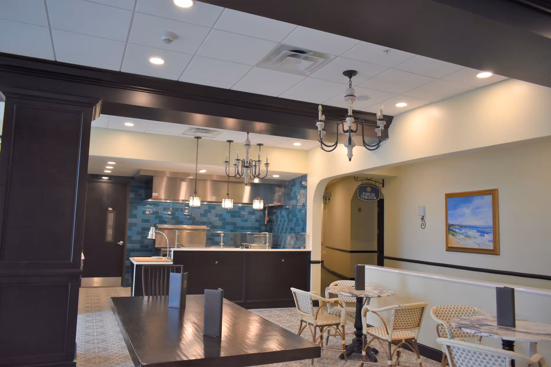 Interior view of a dining area in a senior living facility featuring a long dark wooden table with menus, smaller round tables with wicker chairs, a kitchen area with blue tiled backsplash and stainless steel appliances, chandeliers hanging from the ceiling, and a hallway with a sign for the Forum Theater.