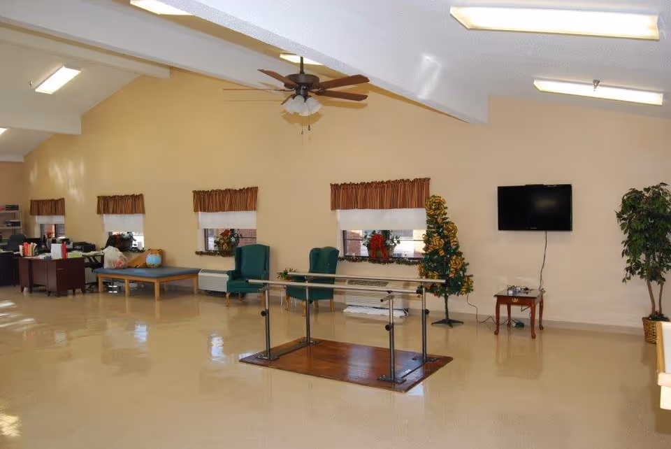 A spacious room with beige walls and a shiny floor, featuring a ceiling fan with lights, three small windows with brown valances, two green chairs, a small decorated artificial tree, a wall-mounted flat-screen TV above a small wooden table, and a parallel bar setup in the center of the room. There is also a desk area with office supplies and a globe on a table in the background.