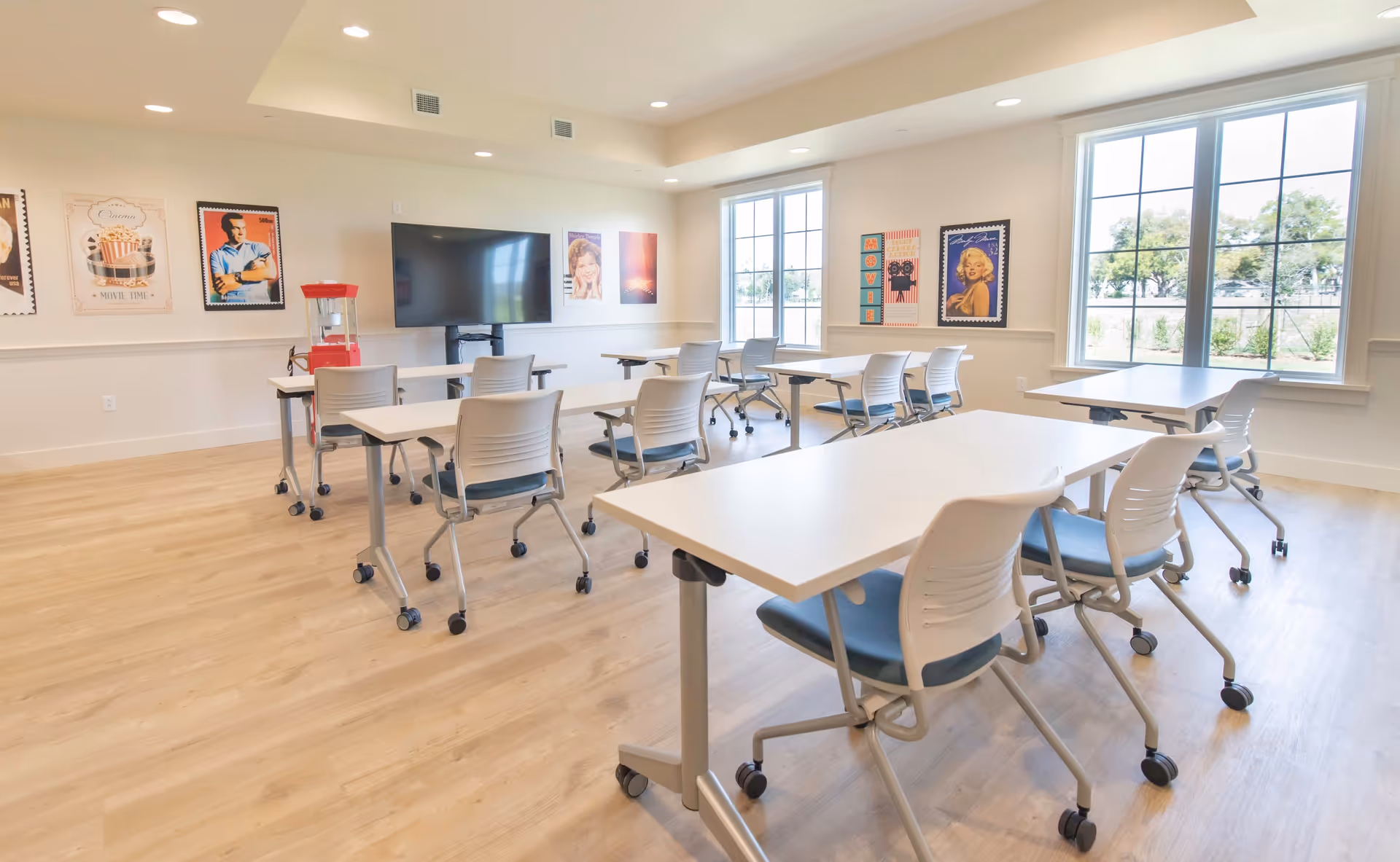 Bright multipurpose activity room with white rolling tables and chairs, a large wall TV, popcorn machine and framed posters by big windows.