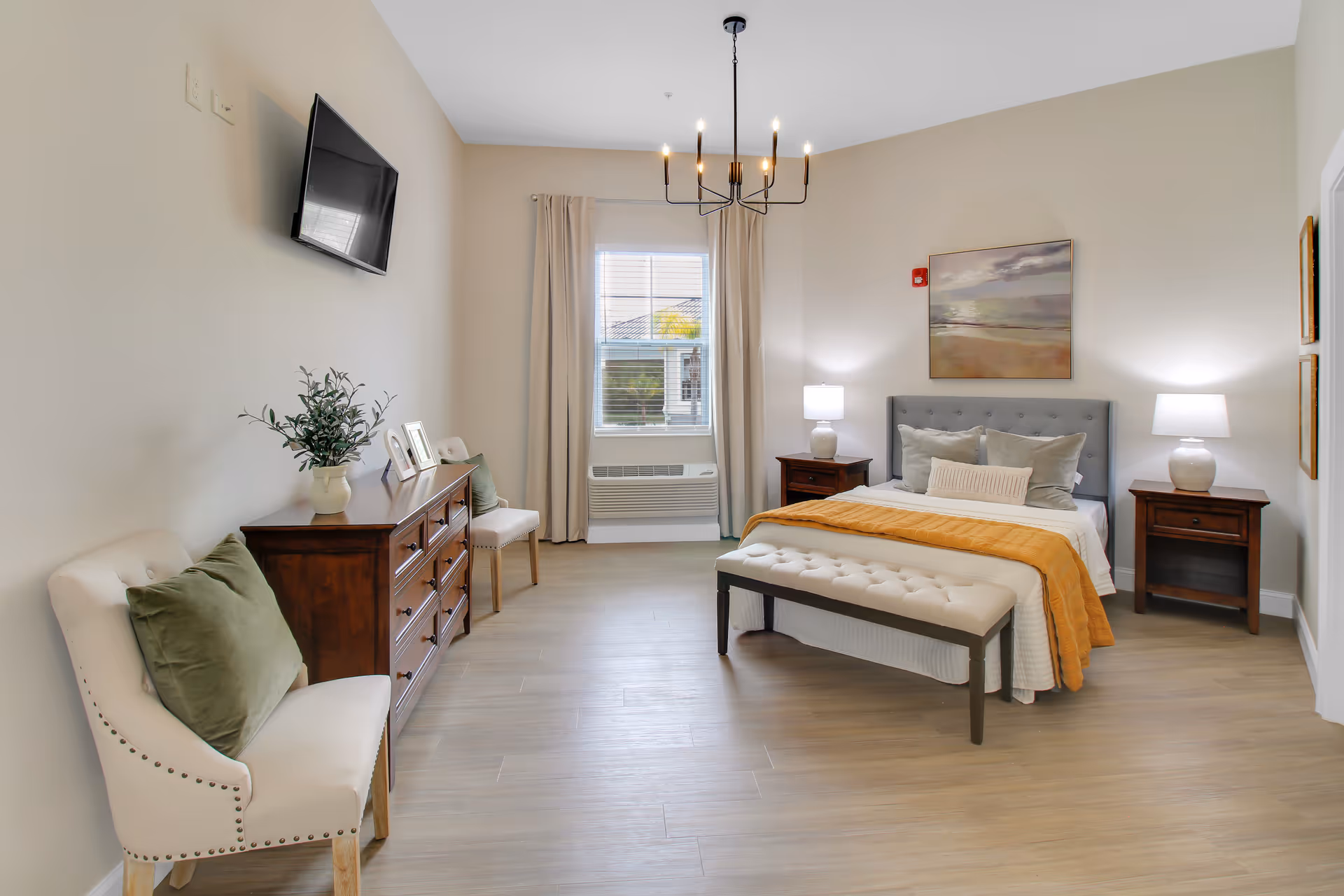 A bright and spacious bedroom with a large bed featuring a tufted headboard, white and gray bedding, and an orange throw blanket. There are two wooden nightstands with white lamps on either side of the bed. A cushioned bench sits at the foot of the bed. The room has light-colored walls and wood flooring, a window with beige curtains, a wooden dresser with decorative items, two white chairs with green pillows, and a flat-screen TV mounted on the wall. A modern chandelier hangs from the ceiling.