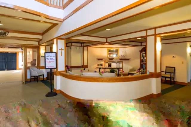 Interior view of a senior living facility reception area with a curved wooden front desk, seating, and warm lighting.