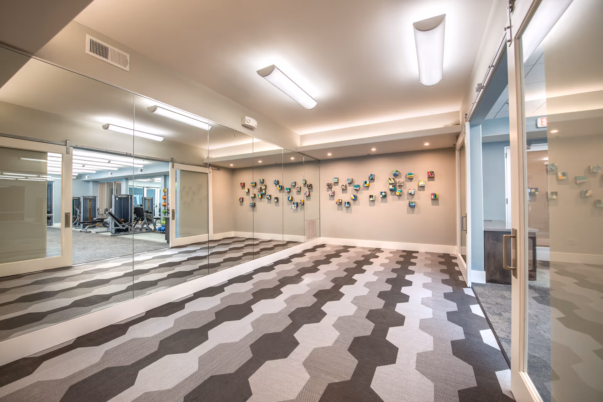 Empty mirrored exercise/stretch room with geometric patterned carpet and a view into an adjacent fitness area.