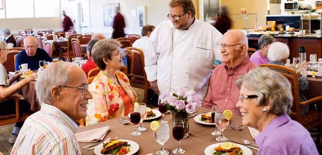 A group of elderly people sitting around a dining table in a senior living facility, enjoying a meal and conversing with a chef standing beside them. The table is set with plates of food, glasses of water with lemon, and a vase with flowers. Other residents and staff are visible in the background in a bright, spacious dining room.