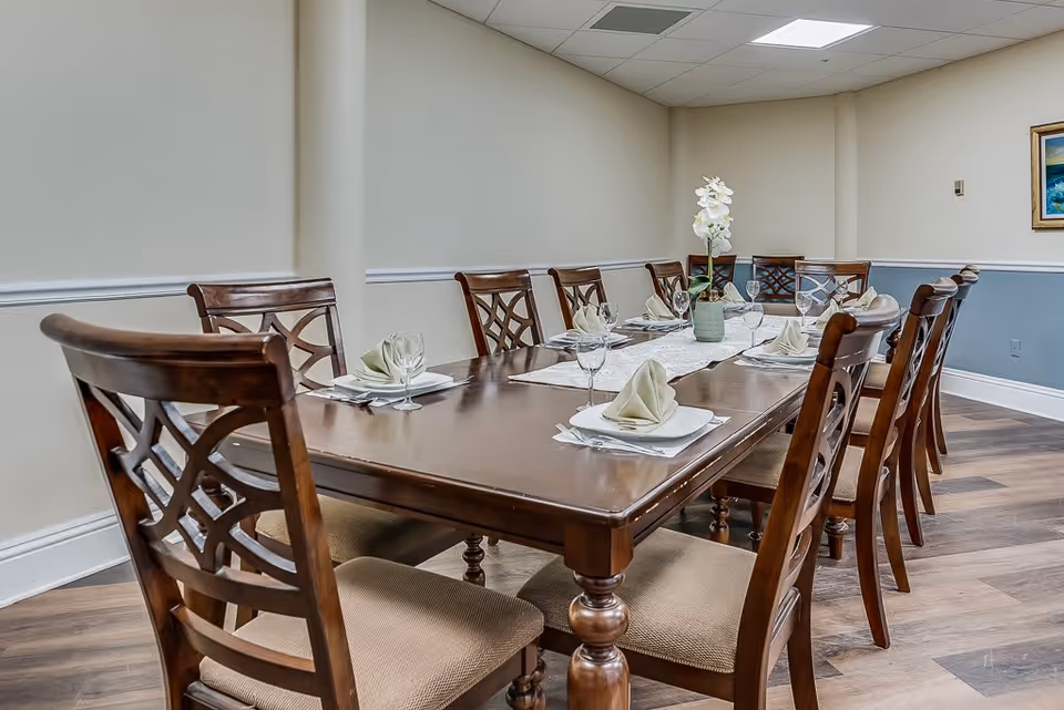 A long wooden dining table set with eight chairs, each place setting includes a white plate, a folded beige napkin, and a wine glass. A white orchid centerpiece in a green vase is placed in the middle of the table. The room has light-colored walls with a blue wainscoting and a framed painting on the wall.