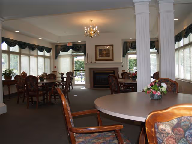 A spacious dining room with multiple round tables and cushioned chairs featuring floral upholstery. The room has large windows with green valances allowing natural light to enter. There is a fireplace centered on the far wall with a framed picture above it, and a chandelier hanging from the ceiling. Decorative columns are also visible in the room.